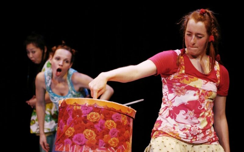 Drei Frauen auf der Bühne; eine vorne steckt ihre Hand in eine verzierte Trommel, während zwei andere im Hintergrund mit überraschten Gesichtern zuschauen.