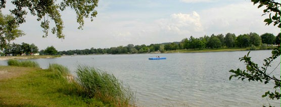 Ein ruhiger See, gesäumt von Gras und Bäumen, in dessen Mitte unter einem teilweise bewölkten Himmel ein kleines blaues Boot schwimmt.