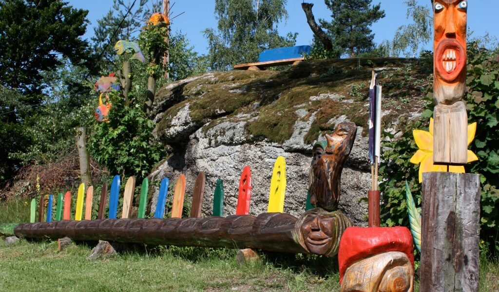 Eine Reihe bunter Totempfähle aus Holz und geschnitzte Gesichter, die im Freien auf Gras in der Nähe eines felsigen Hügels mit Bäumen und blauem Himmel im Hintergrund ausgestellt sind.