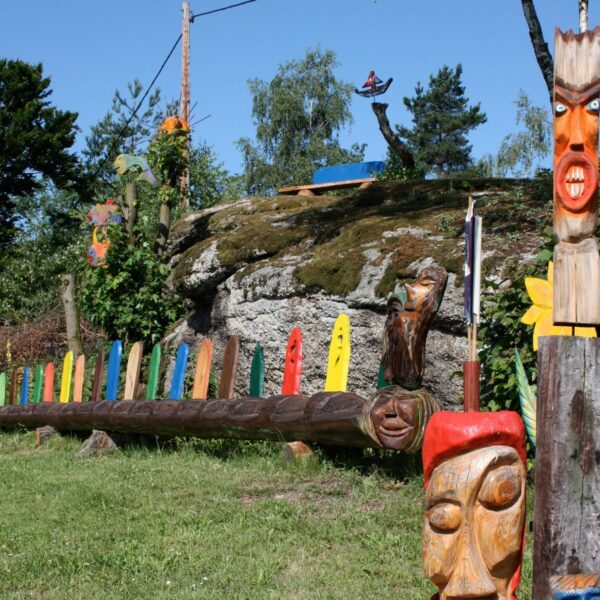 Eine Reihe bunter Totempfähle aus Holz und geschnitzte Gesichter, die im Freien auf Gras in der Nähe eines felsigen Hügels mit Bäumen und blauem Himmel im Hintergrund ausgestellt sind.