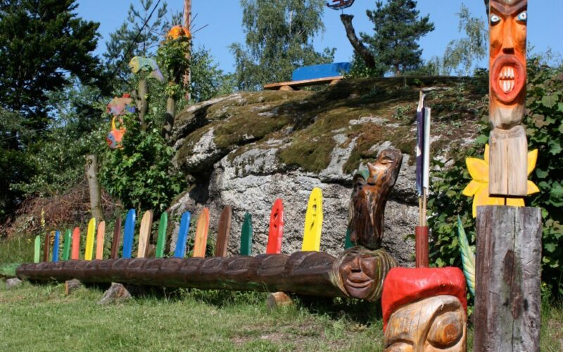Eine Reihe bunter Totempfähle aus Holz und geschnitzte Gesichter, die im Freien auf Gras in der Nähe eines felsigen Hügels mit Bäumen und blauem Himmel im Hintergrund ausgestellt sind.