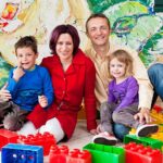 Eine vierköpfige Familie sitzt auf dem Boden, umgeben von großen bunten Spielzeugklötzen, und lächelt in einem hell dekorierten Innenraum in die Kamera.