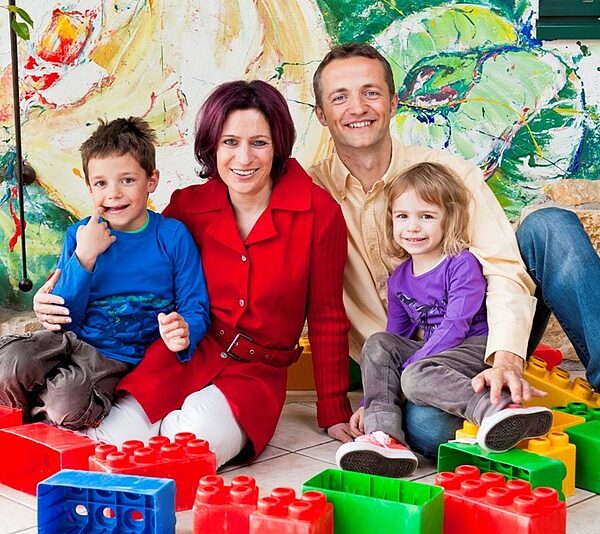 Eine vierköpfige Familie sitzt auf dem Boden, umgeben von großen bunten Spielzeugklötzen, und lächelt in einem hell dekorierten Innenraum in die Kamera.