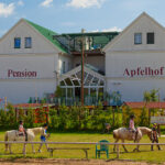 Zwei Kinder reiten an einem sonnigen Tag auf Pferden vor einem weißen Gebäude mit der Aufschrift "Pension Apfelhof", im Hintergrund sind Grünflächen und Spielgeräte zu sehen.