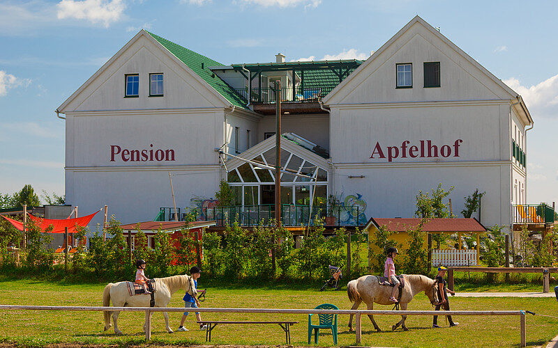 Zwei Kinder reiten an einem sonnigen Tag auf Pferden vor einem weißen Gebäude mit der Aufschrift "Pension Apfelhof", im Hintergrund sind Grünflächen und Spielgeräte zu sehen.