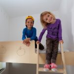 Zwei kleine Kinder, eines in Blau und eines in Lila, lächeln, während sie auf einer hölzernen Etagenbettleiter in einem weißen Raum sitzen und stehen.