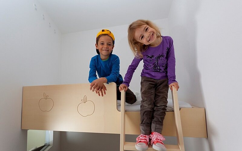 Zwei kleine Kinder, eines in Blau und eines in Lila, lächeln, während sie auf einer hölzernen Etagenbettleiter in einem weißen Raum sitzen und stehen.