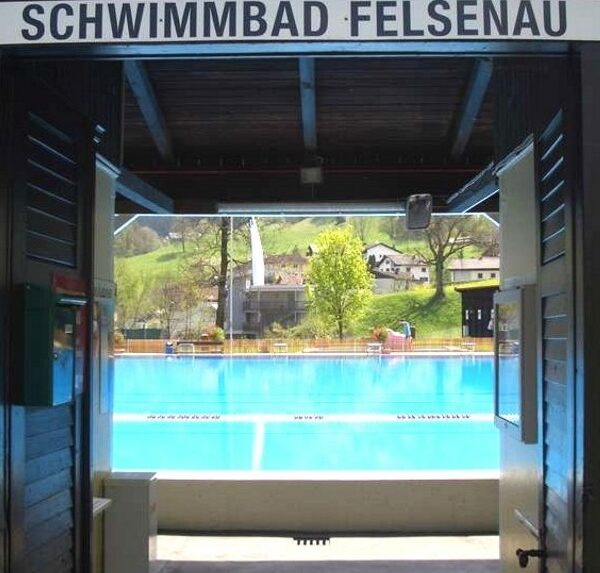 Eingang zum Schwimmbad Felsenau mit einer offenen Tür, die zu einem Freibad führt, mit Blick auf die Hügel und Häuser im Hintergrund.
