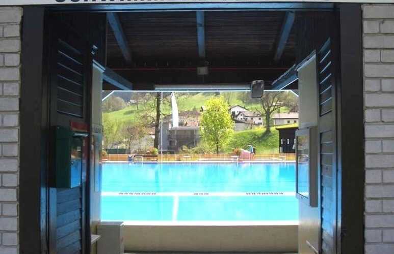 Eingang zum Schwimmbad Felsenau mit einer offenen Tür, die zu einem Freibad führt, mit Blick auf die Hügel und Häuser im Hintergrund.