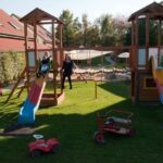 Zwei Kinder spielen auf einem Holzspielplatz mit Rutschen und Schaukeln, umgeben von Gras. Im Vordergrund sind Spielzeugfahrzeuge auf dem Boden verstreut.