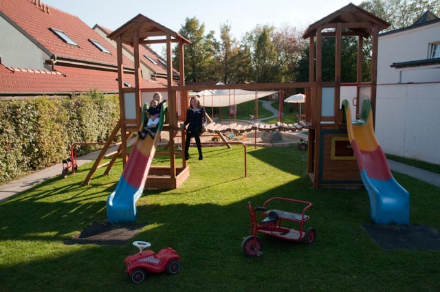 Zwei Kinder spielen auf einem Holzspielplatz mit Rutschen und Schaukeln, umgeben von Gras. Im Vordergrund sind Spielzeugfahrzeuge auf dem Boden verstreut.