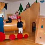 Drei Kinder spielen in einem Innenraum mit einer Holzeisenbahn und einer Kletterwand, umgeben von bemalten Baumdekorationen an den Wänden.