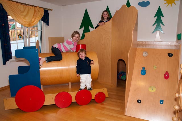 Drei Kinder spielen in einem Innenraum mit einer Holzeisenbahn und einer Kletterwand, umgeben von bemalten Baumdekorationen an den Wänden.