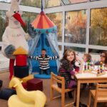 Kinder spielen in einem hell erleuchteten Raum mit Spielzeug, einem Spielzelt und einer Person in einem Storchenkostüm. Zwei Erwachsene beaufsichtigen sie an einem Tisch mit Perlenlabyrinthen.