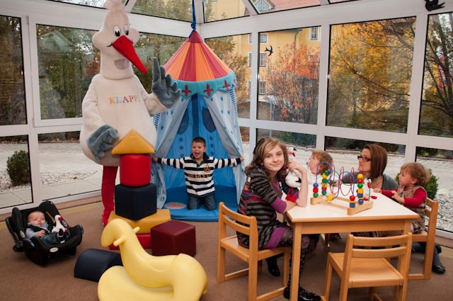 Kinder spielen in einem hell erleuchteten Raum mit Spielzeug, einem Spielzelt und einer Person in einem Storchenkostüm. Zwei Erwachsene beaufsichtigen sie an einem Tisch mit Perlenlabyrinthen.