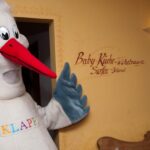 Eine Person in einem Storchenkostüm mit der Aufschrift "KLAPPE" steht in einem Innenraum neben einer Wand mit der Aufschrift "Baby-Küche, Wickelraum, Surfen, Internet".