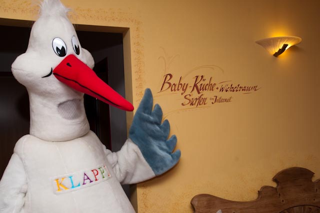 Eine Person in einem Storchenkostüm mit der Aufschrift "KLAPPE" steht in einem Innenraum neben einer Wand mit der Aufschrift "Baby-Küche, Wickelraum, Surfen, Internet".