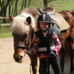 Ein Kind, das einen Helm und eine Weste trägt, steht an einem sonnigen Tag neben einem braunen Pony mit einem Sattel. Eine weitere Person ist teilweise im Hintergrund zu sehen.
