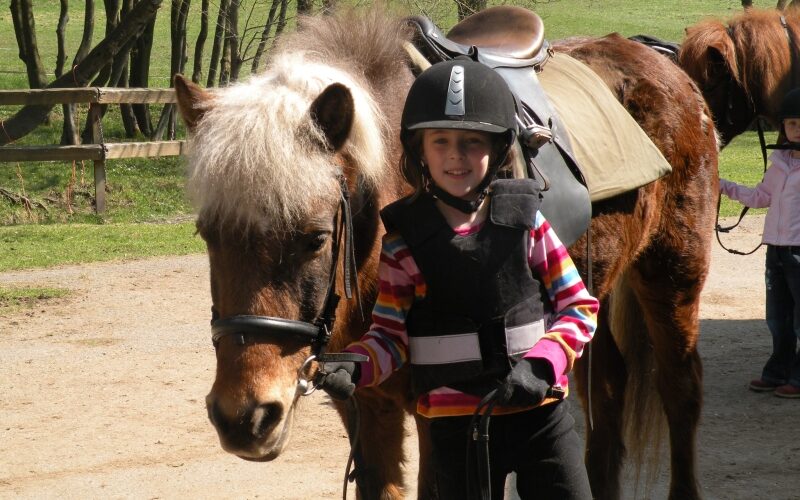Ein Kind, das einen Helm und eine Weste trägt, steht an einem sonnigen Tag neben einem braunen Pony mit einem Sattel. Eine weitere Person ist teilweise im Hintergrund zu sehen.
