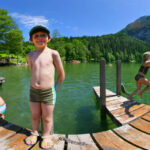 Kinder in Badeanzügen spielen und schwimmen an einem Holzsteg auf einem See, der von grünen Hügeln und Bergen unter einem klaren blauen Himmel umgeben ist.