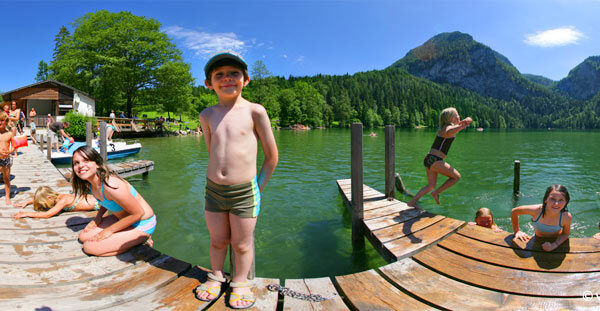 Kinder in Badeanzügen spielen und schwimmen an einem Holzsteg auf einem See, der von grünen Hügeln und Bergen unter einem klaren blauen Himmel umgeben ist.
