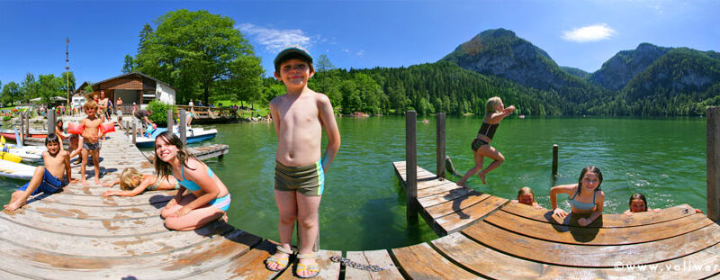 Kinder in Badeanzügen spielen und schwimmen an einem Holzsteg auf einem See, der von grünen Hügeln und Bergen unter einem klaren blauen Himmel umgeben ist.