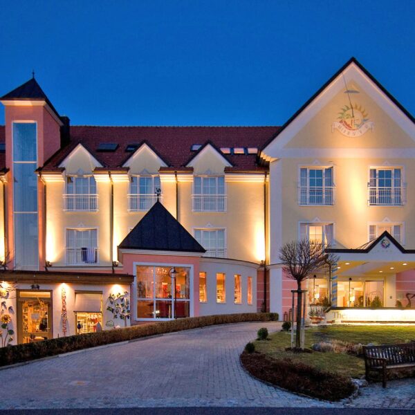 Ein großes, gut beleuchtetes Hotelgebäude mit vielen Fenstern und einem roten Dach, fotografiert in der Abenddämmerung, mit einer Einfahrt und einem angelegten Garten davor.