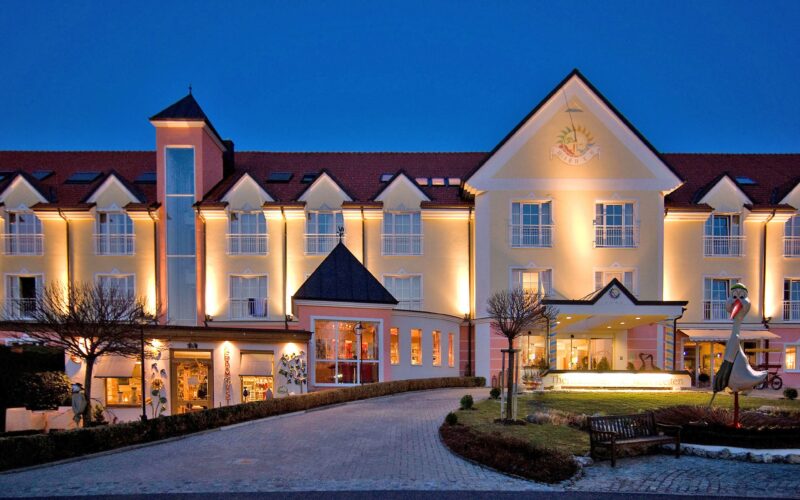 Ein großes, gut beleuchtetes Hotelgebäude mit vielen Fenstern und einem roten Dach, fotografiert in der Abenddämmerung, mit einer Einfahrt und einem angelegten Garten davor.