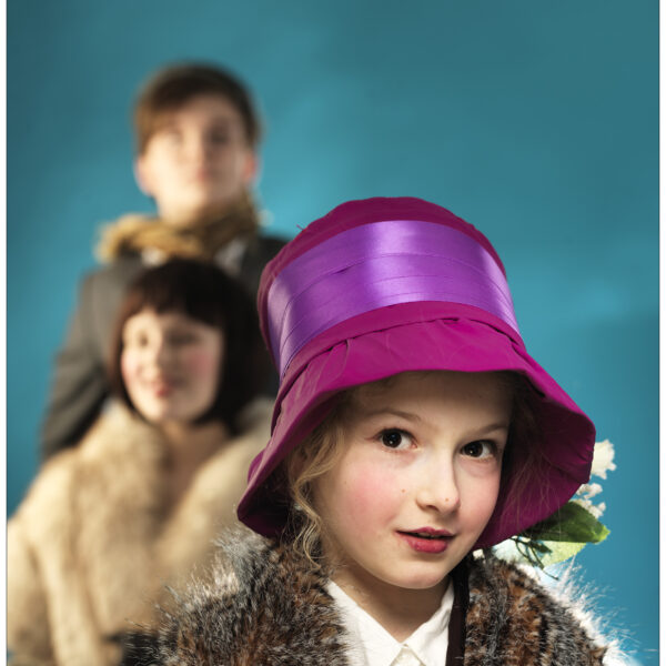 Im Fokus steht ein junges Mädchen mit einem leuchtend lila Hut und einer Pelzstola, im Hintergrund sind vor einem blauen Hintergrund zwei Personen unscharf zu sehen.