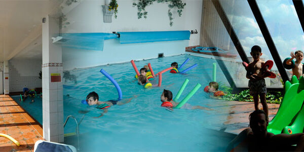 Kinder schwimmen mit Schwimmnudeln in einem Hallenbad mit großen Glasfenstern. Andere stehen in einer Reihe neben einer grünen Rutsche und warten auf ihren Einsatz.