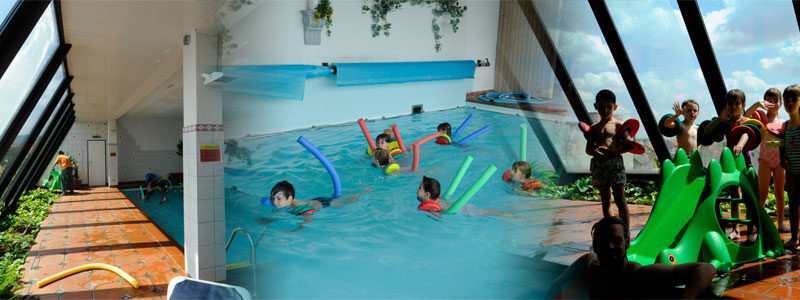Kinder schwimmen mit Schwimmnudeln in einem Hallenbad mit großen Glasfenstern. Andere stehen in einer Reihe neben einer grünen Rutsche und warten auf ihren Einsatz.