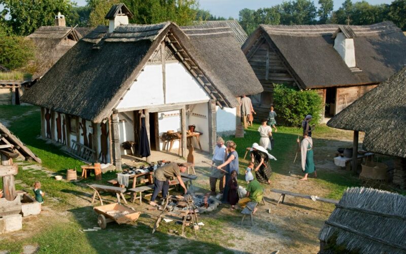 Menschen in historischen Kostümen versammeln sich um strohgedeckte Gebäude und widmen sich in einer rekonstruierten Dorfkulisse Outdoor-Aktivitäten und Kunsthandwerk.