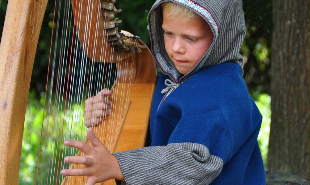 Ein kleines Kind in einem blauen Kapuzenmantel spielt im Freien eine Holzharfe. Dabei konzentriert es sich mit einer Hand auf die Saiten und hält mit der anderen das Instrument.