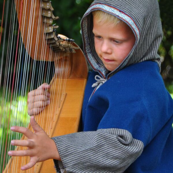 Ein kleines Kind in einem blauen Kapuzenmantel spielt im Freien eine Holzharfe. Dabei konzentriert es sich mit einer Hand auf die Saiten und hält mit der anderen das Instrument.