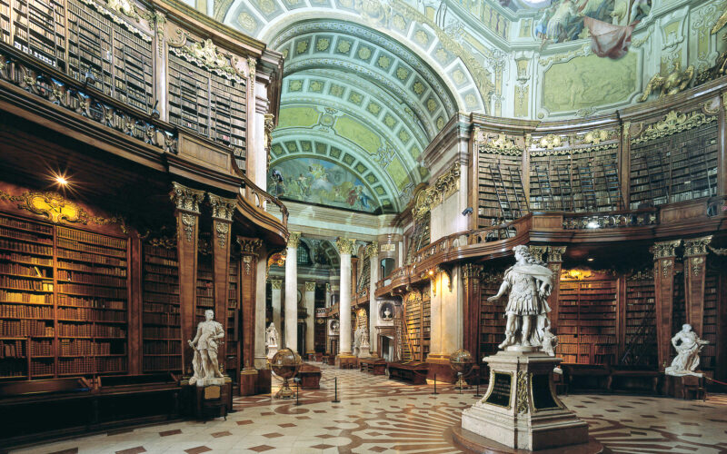 Große, verzierte Bibliothek mit hohen, gewölbten Decken, detaillierten Fresken, hohen Bücherregalen, Marmorstatuen und einem gemusterten Fußboden.