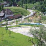 Ein kleiner Park mit Spielplatz, Sandkasten und Spazierwegen ist umgeben von Wiesen, Bäumen und nahegelegenen Wohnhäusern in dörflicher Umgebung.