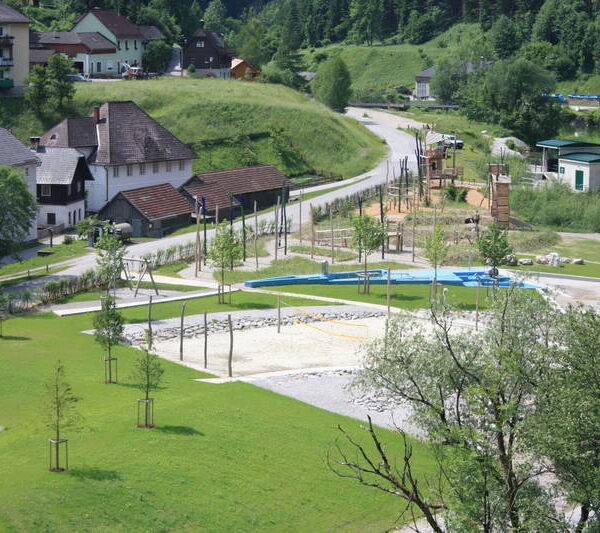 Ein kleiner Park mit Spielplatz, Sandkasten und Spazierwegen ist umgeben von Wiesen, Bäumen und nahegelegenen Wohnhäusern in dörflicher Umgebung.