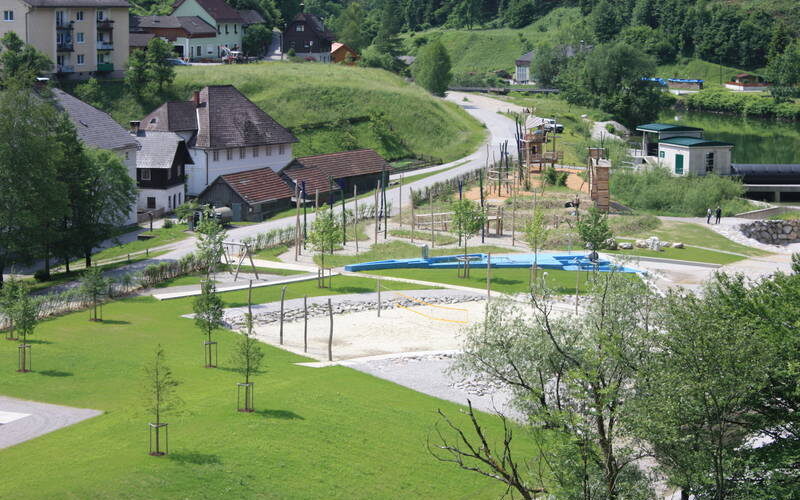 Ein kleiner Park mit Spielplatz, Sandkasten und Spazierwegen ist umgeben von Wiesen, Bäumen und nahegelegenen Wohnhäusern in dörflicher Umgebung.