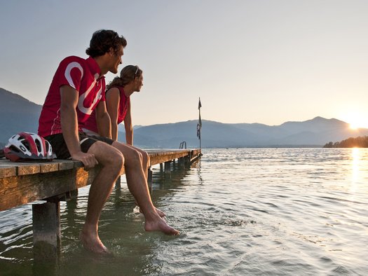 Zwei Menschen sitzen mit den Füßen im Wasser auf einem Holzsteg und beobachten den Sonnenuntergang über einem See mit Bergen im Hintergrund.