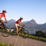 Zwei Menschen mit Helmen und roten Hemden fahren mit dem Fahrrad auf einem gepflasterten Weg, im Hintergrund sind unter einem klaren Himmel Berge, ein See und Grün zu sehen.