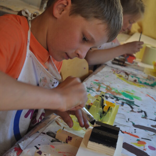Ein Junge in einer Schürze bemalt mit einem Pinsel eine Holzarbeit an einem Tisch, der mit bunten Farbspritzern bedeckt ist. Ein anderes Kind arbeitet im Hintergrund.