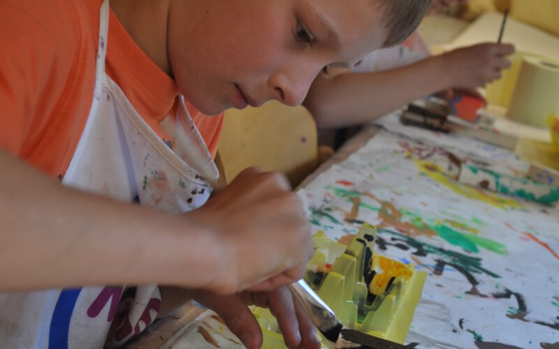 Ein Junge in einer Schürze bemalt mit einem Pinsel eine Holzarbeit an einem Tisch, der mit bunten Farbspritzern bedeckt ist. Ein anderes Kind arbeitet im Hintergrund.