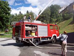 Ein rotes Feuerwehrauto steht mit ausgefahrenem Schlauch auf einer Straße; zwei Personen stehen darauf und eine weitere Person fotografiert die Szene.