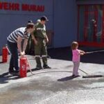 Ein Kind sprüht Wasser aus einem Schlauch, unterstützt von zwei Erwachsenen, in der Nähe von Feuerlöschern vor einem Gebäude mit der Aufschrift „FEUERWEHR MURAU“.