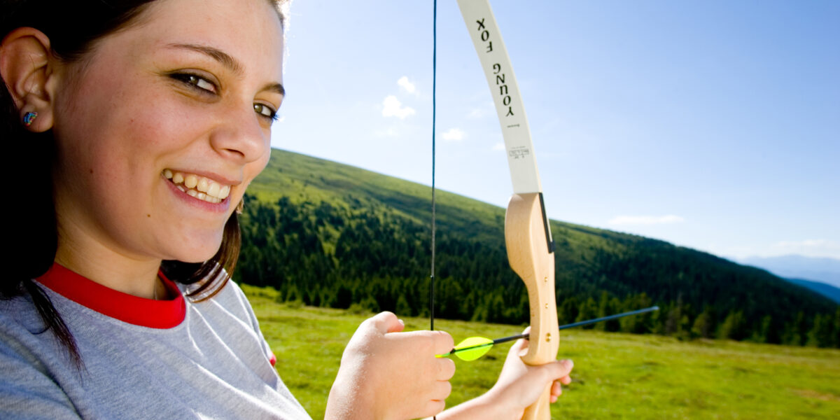 Eine Person, die im Freien lächelt, hält einen Recurve-Bogen mit einem Pfeil in der Hand und steht in einer grasbewachsenen Gegend mit Hügeln und Bäumen im Hintergrund.