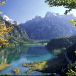 Ein klarer, blauer See, umgeben von bewaldeten Hügeln und hohen, schroffen Bergen unter einem strahlenden Himmel, mit etwas Herbstlaub im Vordergrund.