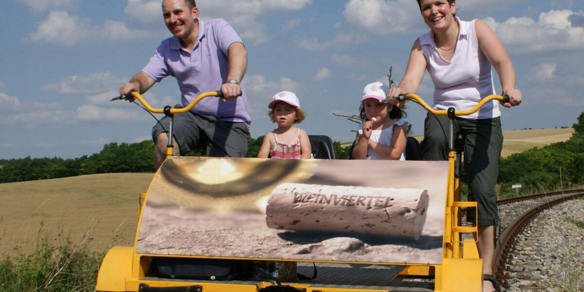 Eine vierköpfige Familie fährt mit einem gelben Schienenfahrrad auf Eisenbahnschienen durch eine ländliche Landschaft unter einem blauen Himmel mit Wolken.