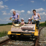 Eine vierköpfige Familie fährt mit einem gelben Schienenfahrrad auf Eisenbahnschienen durch eine ländliche Landschaft unter einem blauen Himmel mit Wolken.