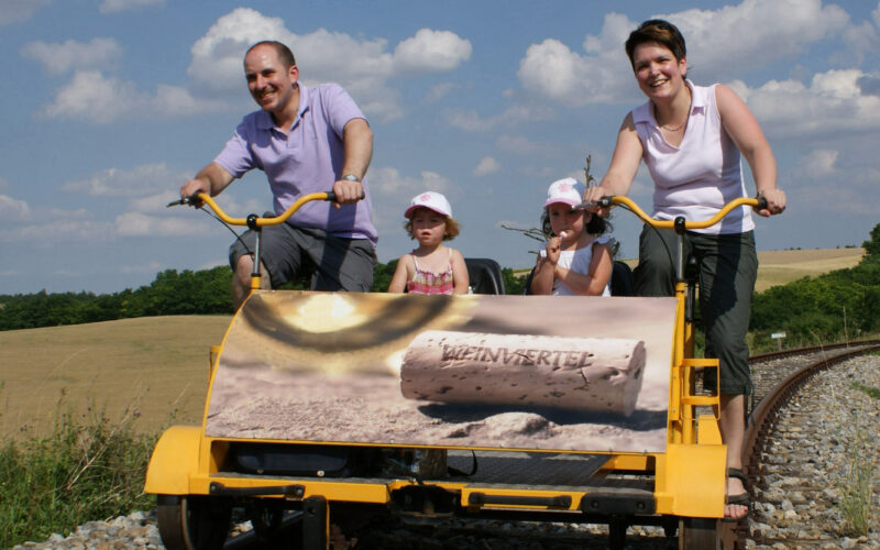 Eine vierköpfige Familie fährt mit einem gelben Schienenfahrrad auf Eisenbahnschienen durch eine ländliche Landschaft unter einem blauen Himmel mit Wolken.