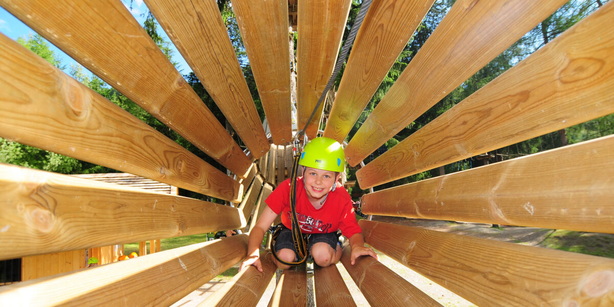 Ein kleines Kind, das einen Helm und einen Klettergurt trägt, krabbelt an einem sonnigen Tag durch eine hölzerne Tunnelstruktur in einem Outdoor-Abenteuerpark.